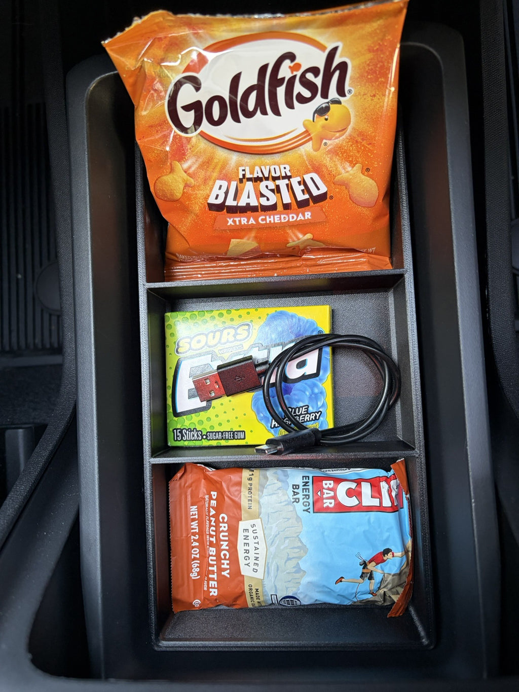 Slide-Out Drawer Tray installed in a Rivian R2 console drawer, top view, holding crackers, gum with a charging cable and an energy bar
